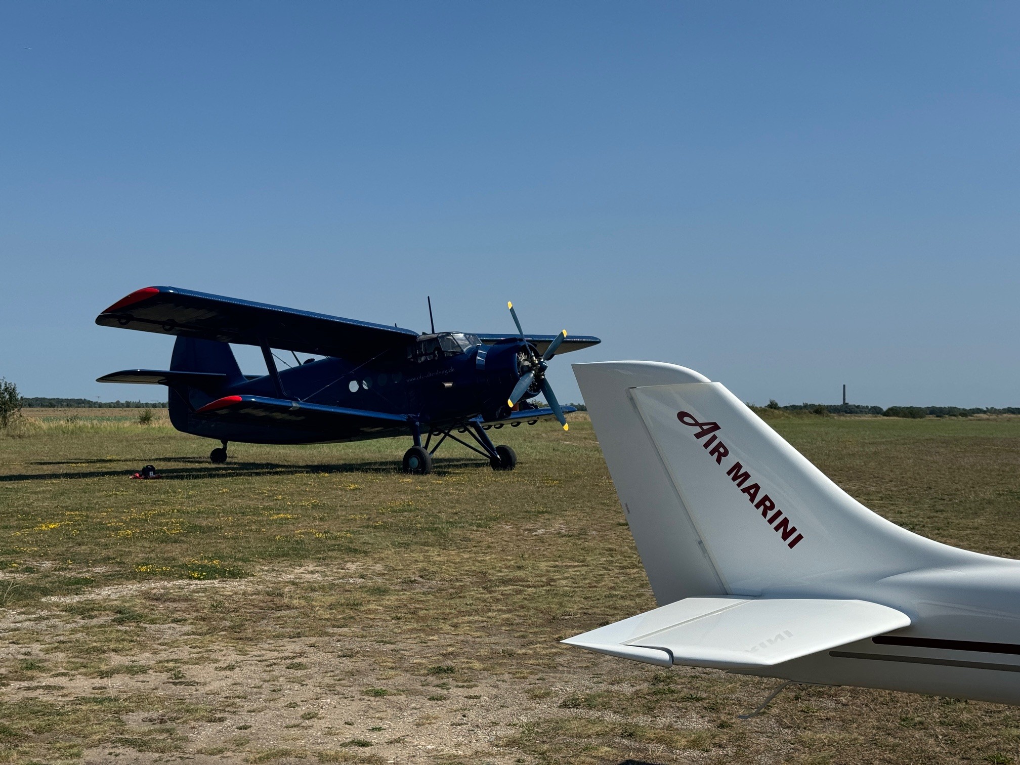 Ein blaues, historisches Doppeldecker-Flugzeug steht neben einem modernen Flugzeug mit Air Marini Schriftzug auf einer Graspiste.