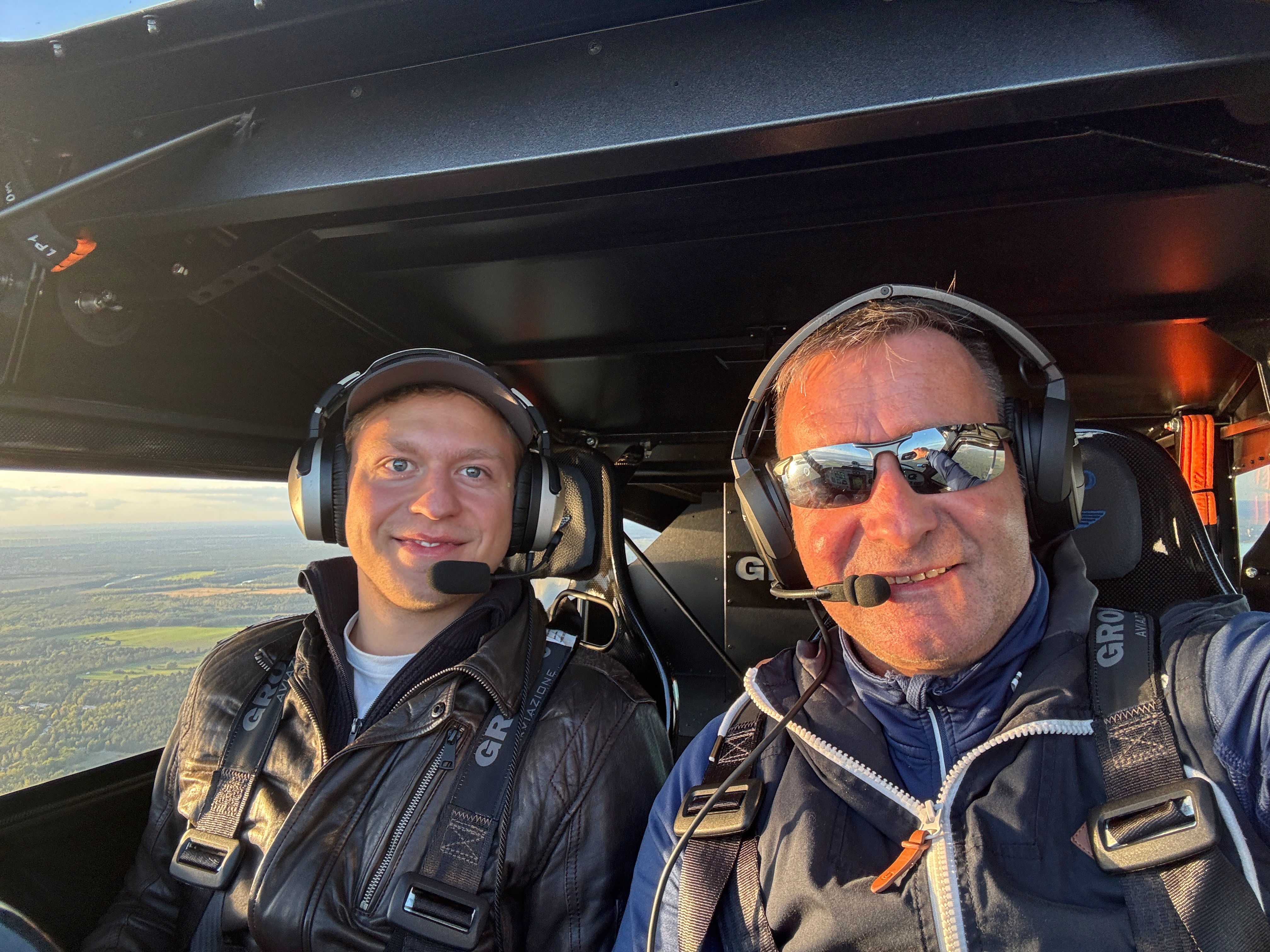 Zwei Männer mit Headsets lächeln in einem Flugzeugcockpit, darunter weite Landschaft im Hintergrund