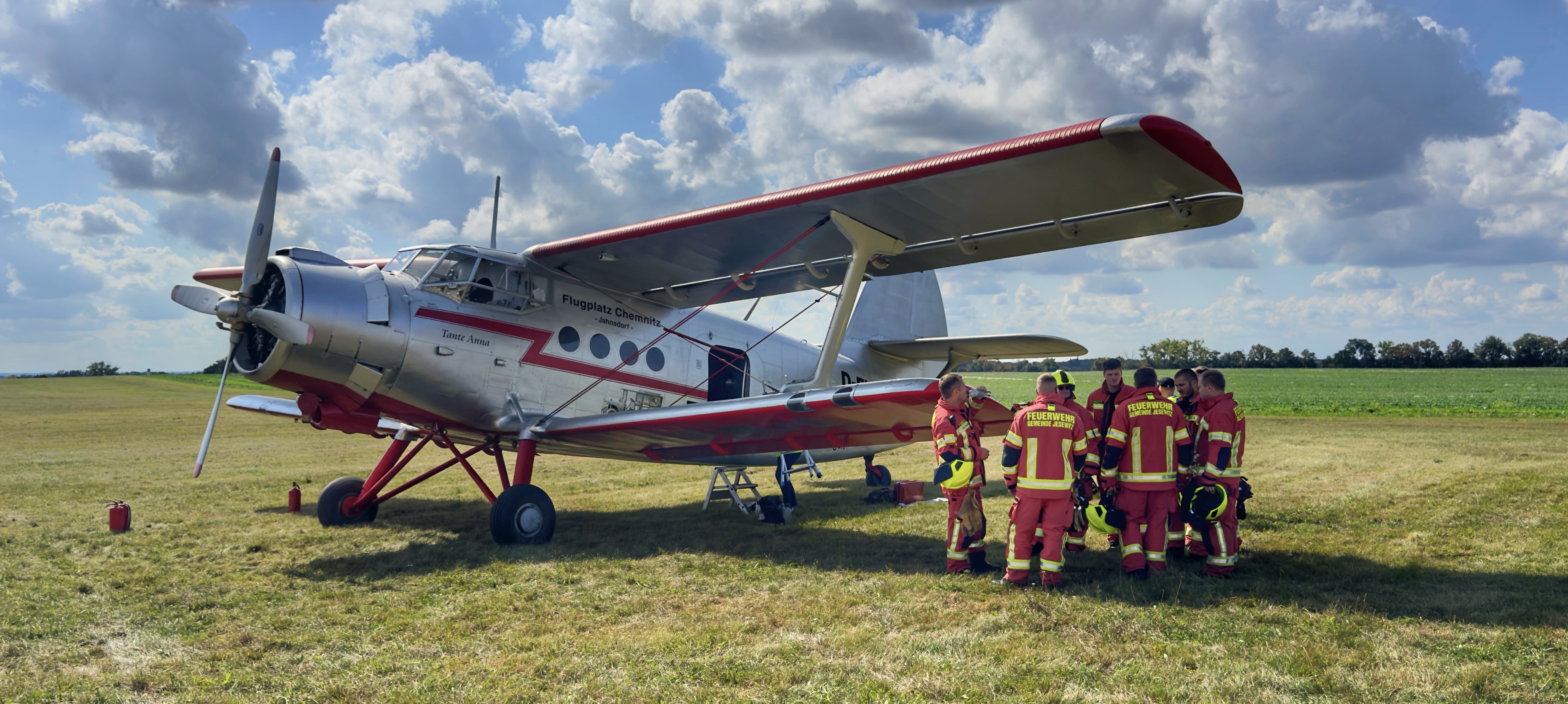 Silbernes Flugzeug mit roter Markierung auf einer Wiese, umgeben von einer Gruppe Feuerwehrleute in Einsatzkleidung, unter blauem Himmel mit Wolken