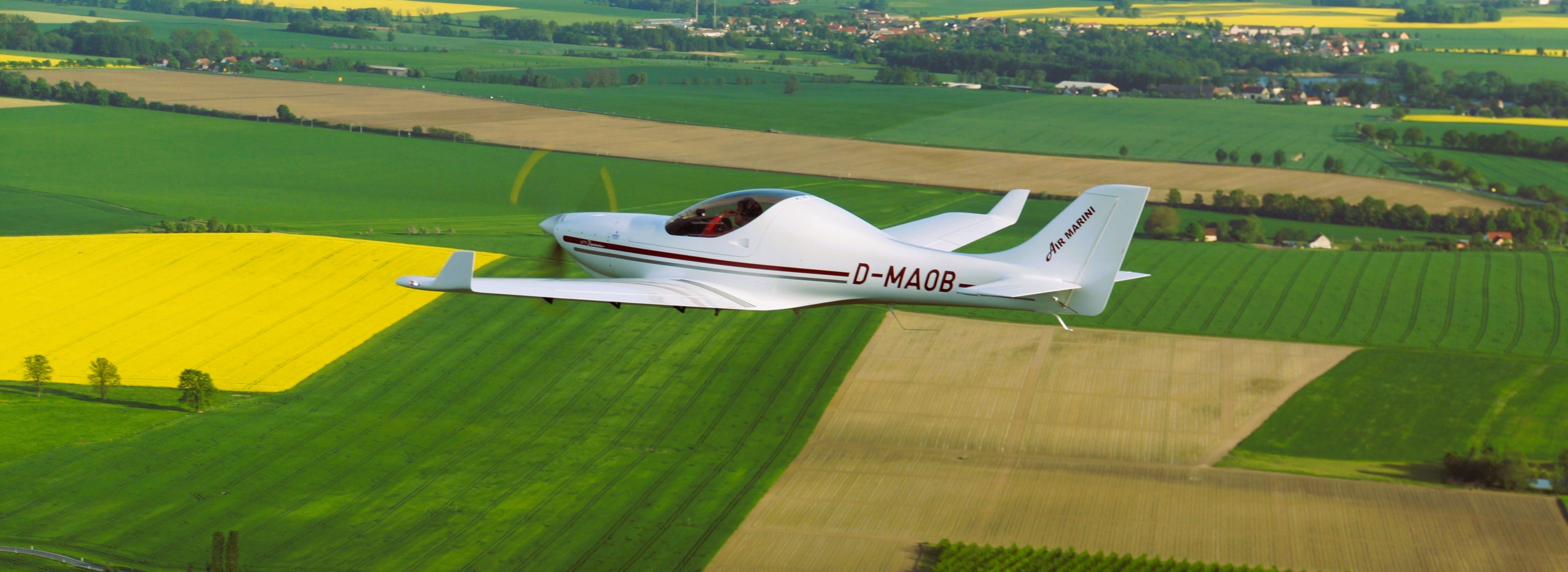 Weißes Leichtflugzeug über grünen und gelben Feldern Ein weißes Leichtflugzeug fliegt über weitläufige Felder, die in verschiedenen Grüntönen und leuchtendem Gelb erstrahlen.