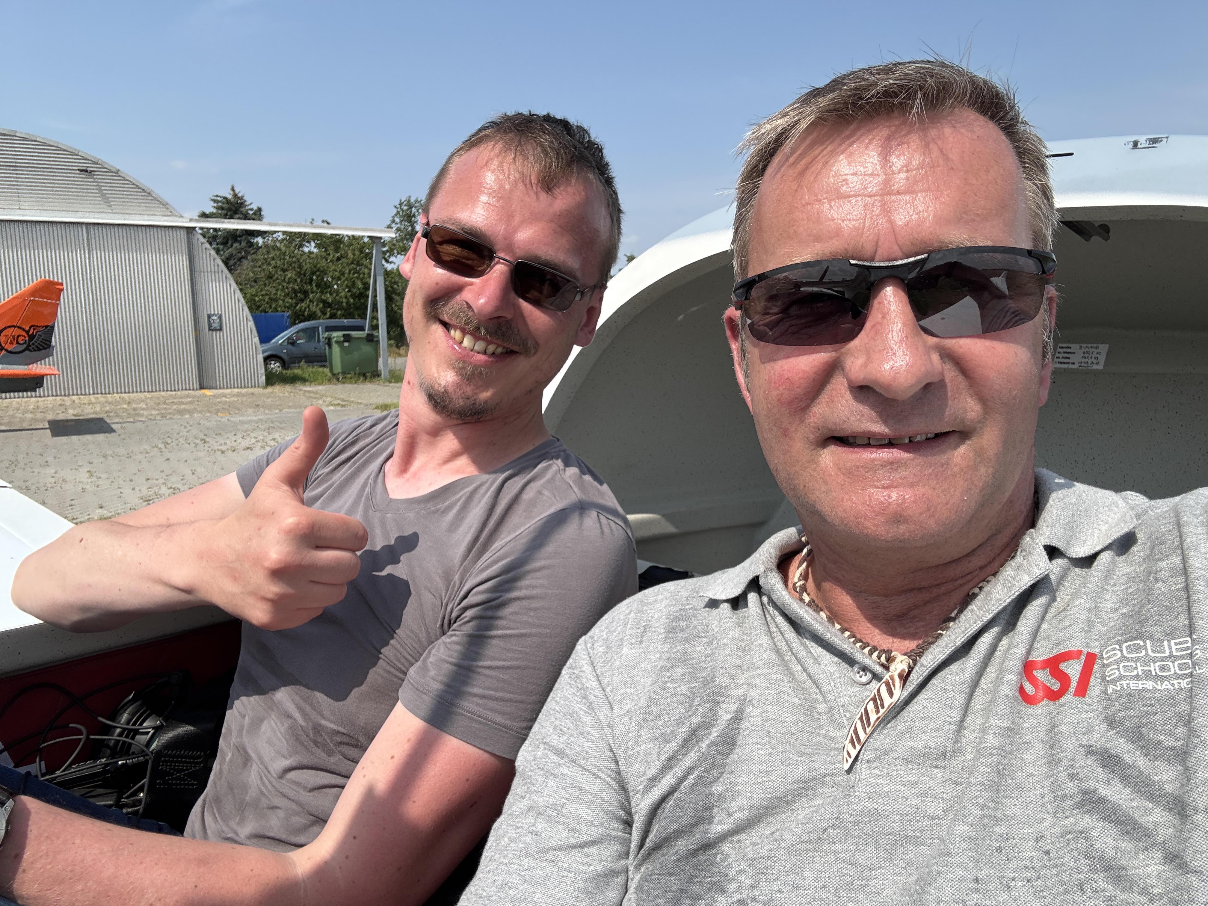 Zwei Männer in einem kleinen Flugzeug, lächelnd und bereit für einen Flug, einer zeigt den Daumen hoch