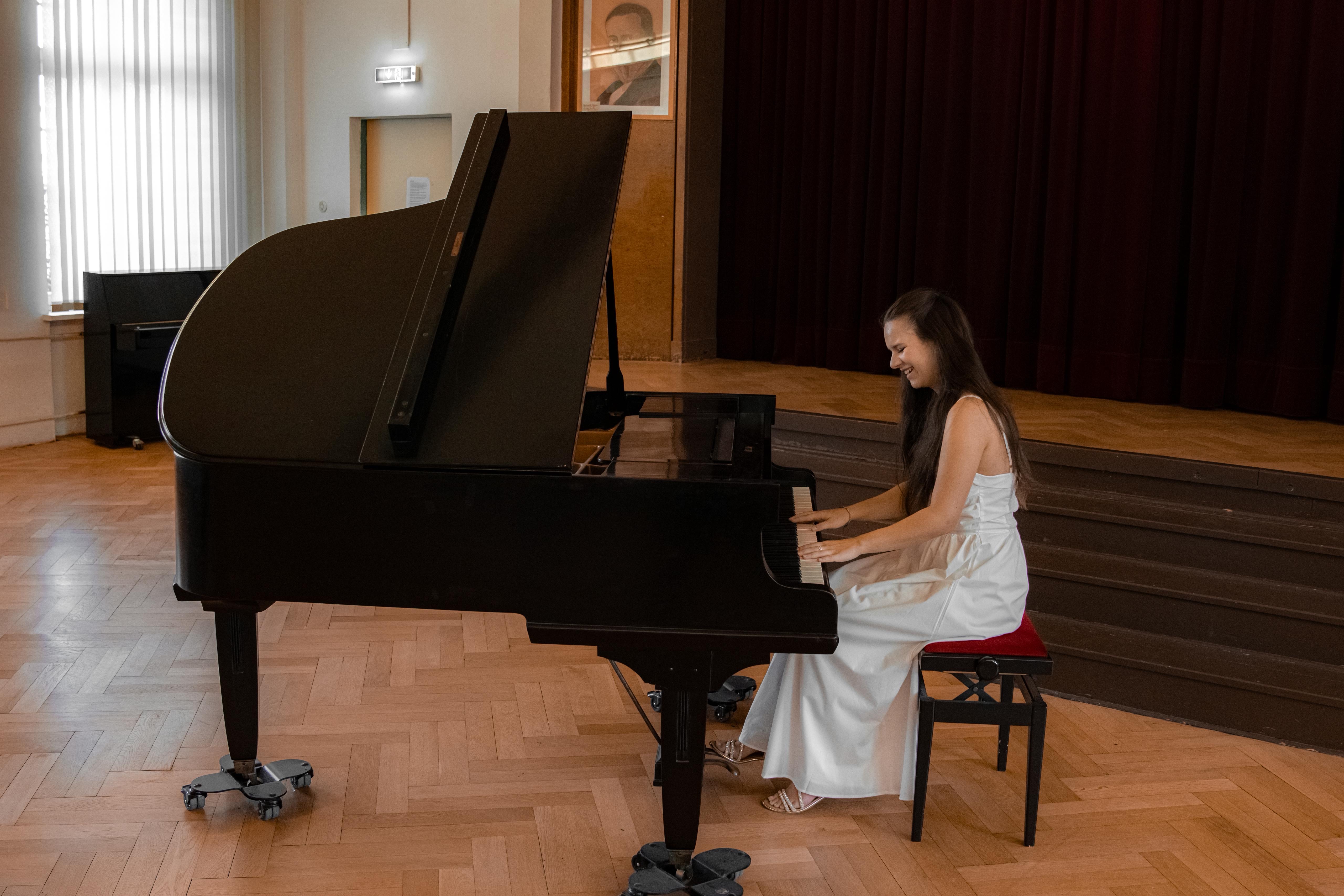 Eine Frau in einem weißen Kleid spielt lächelnd Klavier in einem Konzertsaal mit Holzboden und Bühnenvorhang