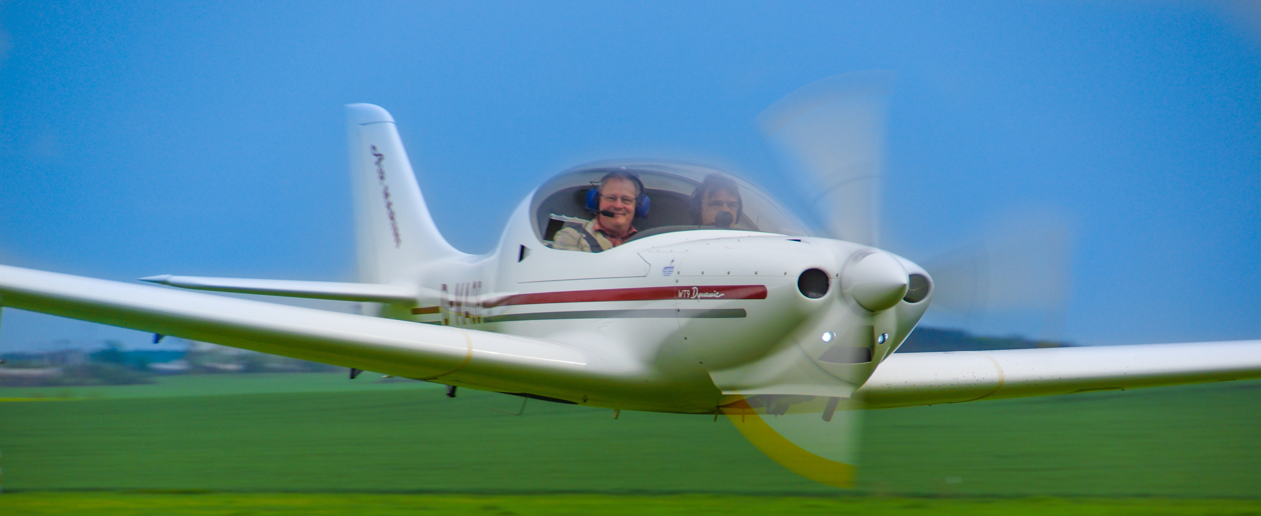 Ein weißes Sportflugzeug mit roter Verzierung fliegt über ein grünes Feld. Im Cockpit sind zwei Personen zu sehen.