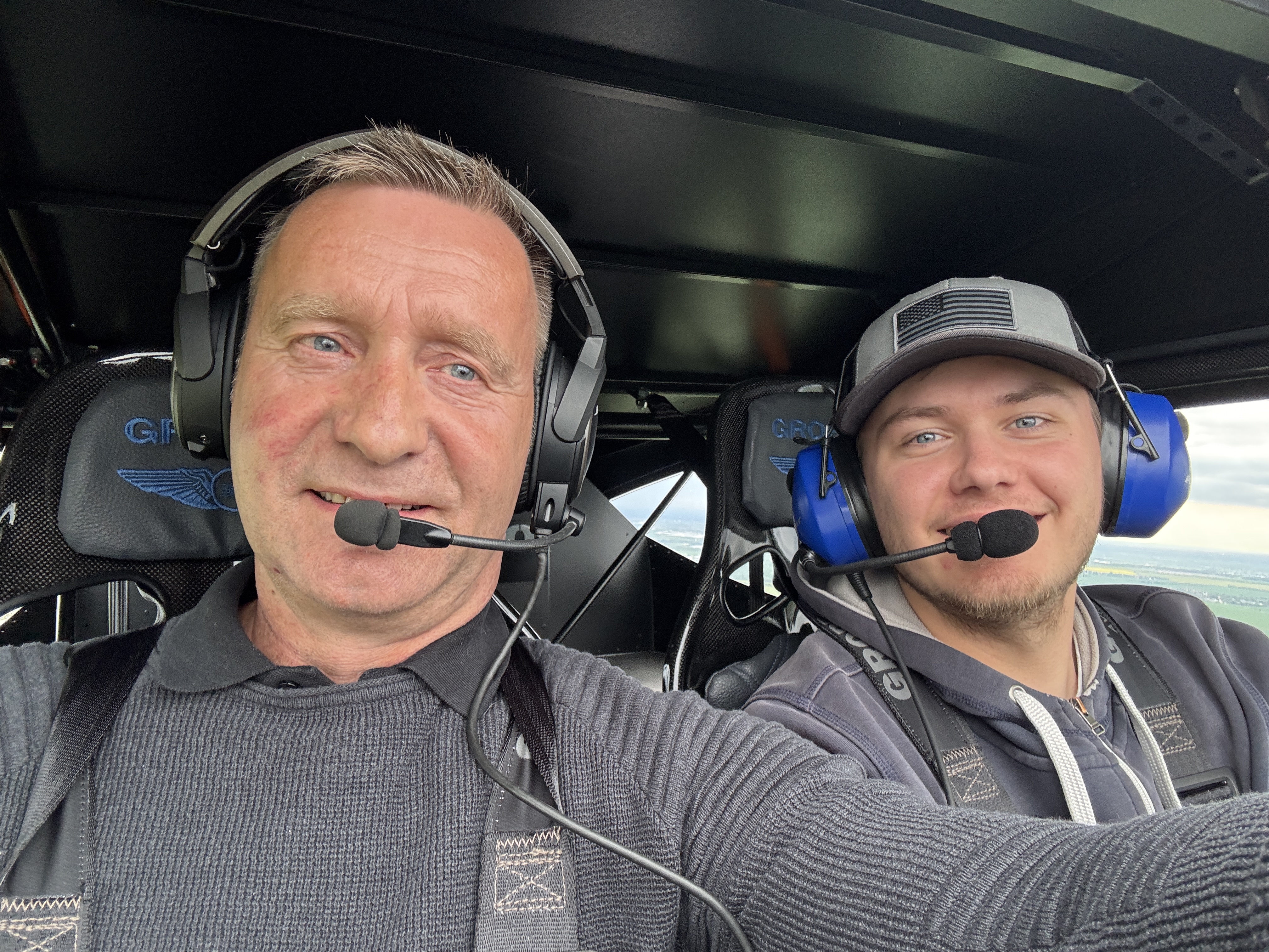 Zwei Männer mit Headsets sitzen in einem Flugzeugcockpit und lächeln in die Kamera