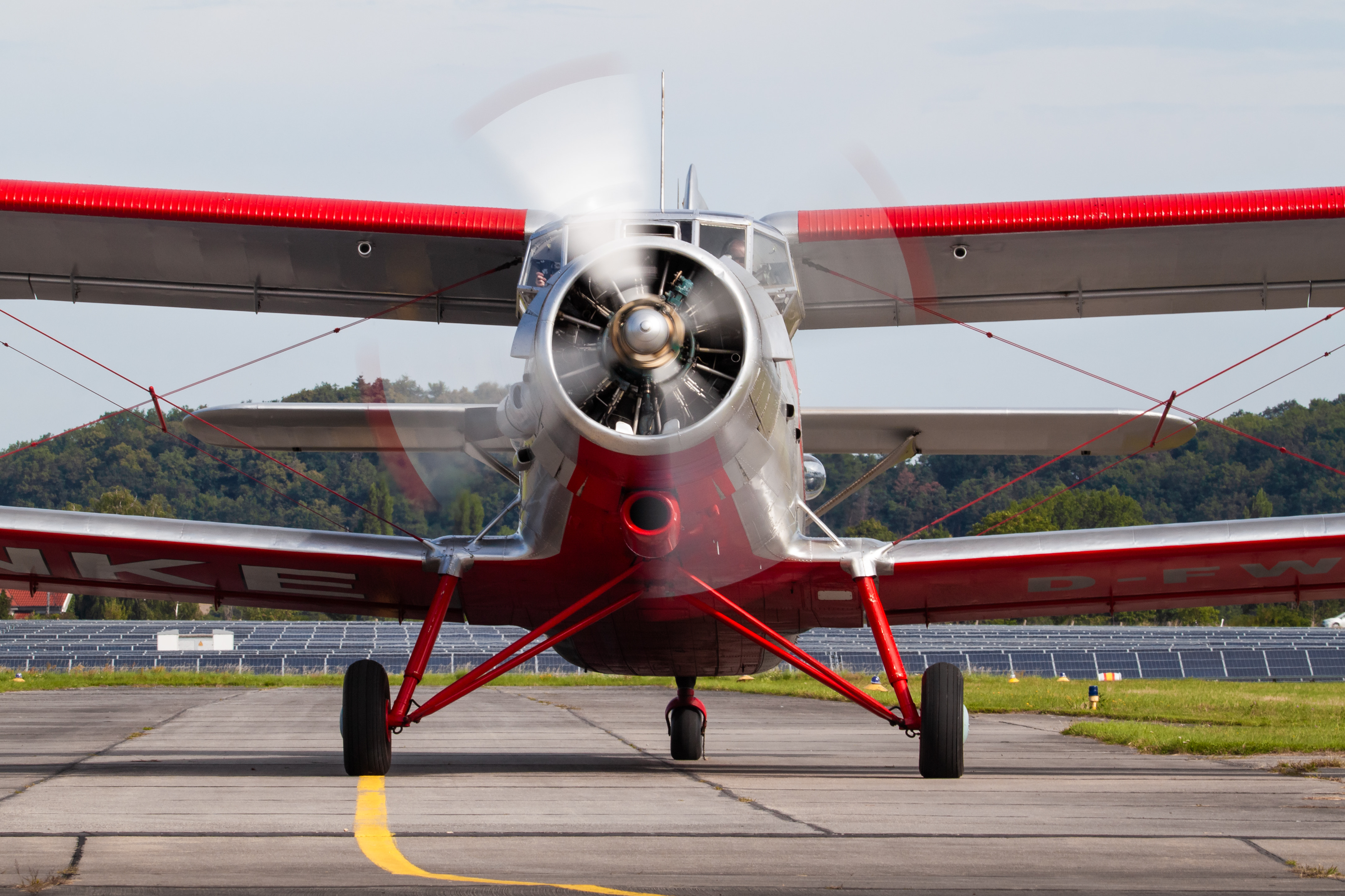 Frontalansicht eines Doppeldeckers mit rot-weißen Akzenten auf dem Rollfeld eines Flugplatzes, umgeben von grüner Landschaft.