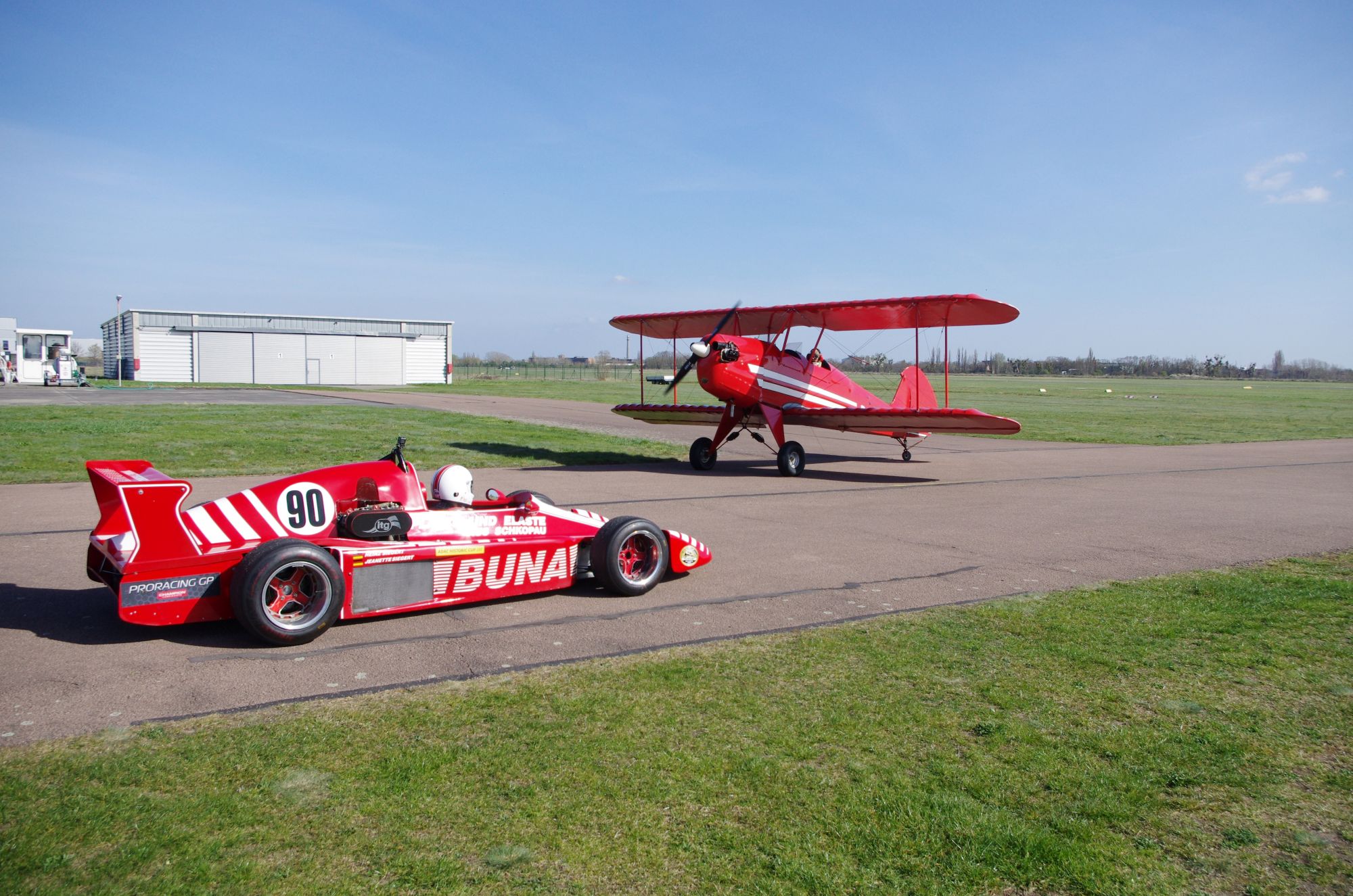 Ein roter Rennwagen neben einem roten Doppeldecker-Flugzeug auf einer Asphaltbahn bei sonnigem Wetter.
