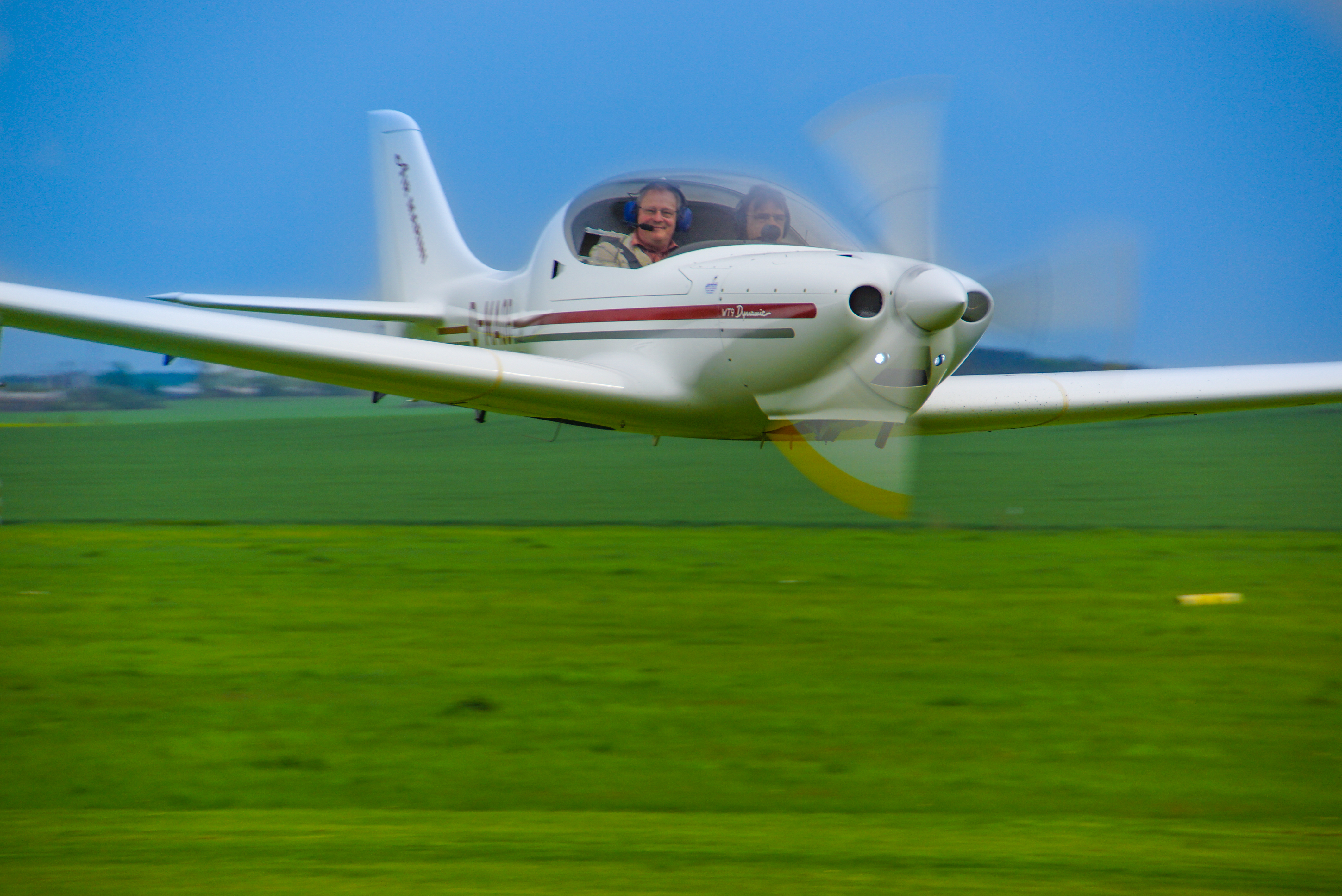 Leichtflugzeug mit zwei Personen in der Kabine fliegt über ein weites grünes Feld
