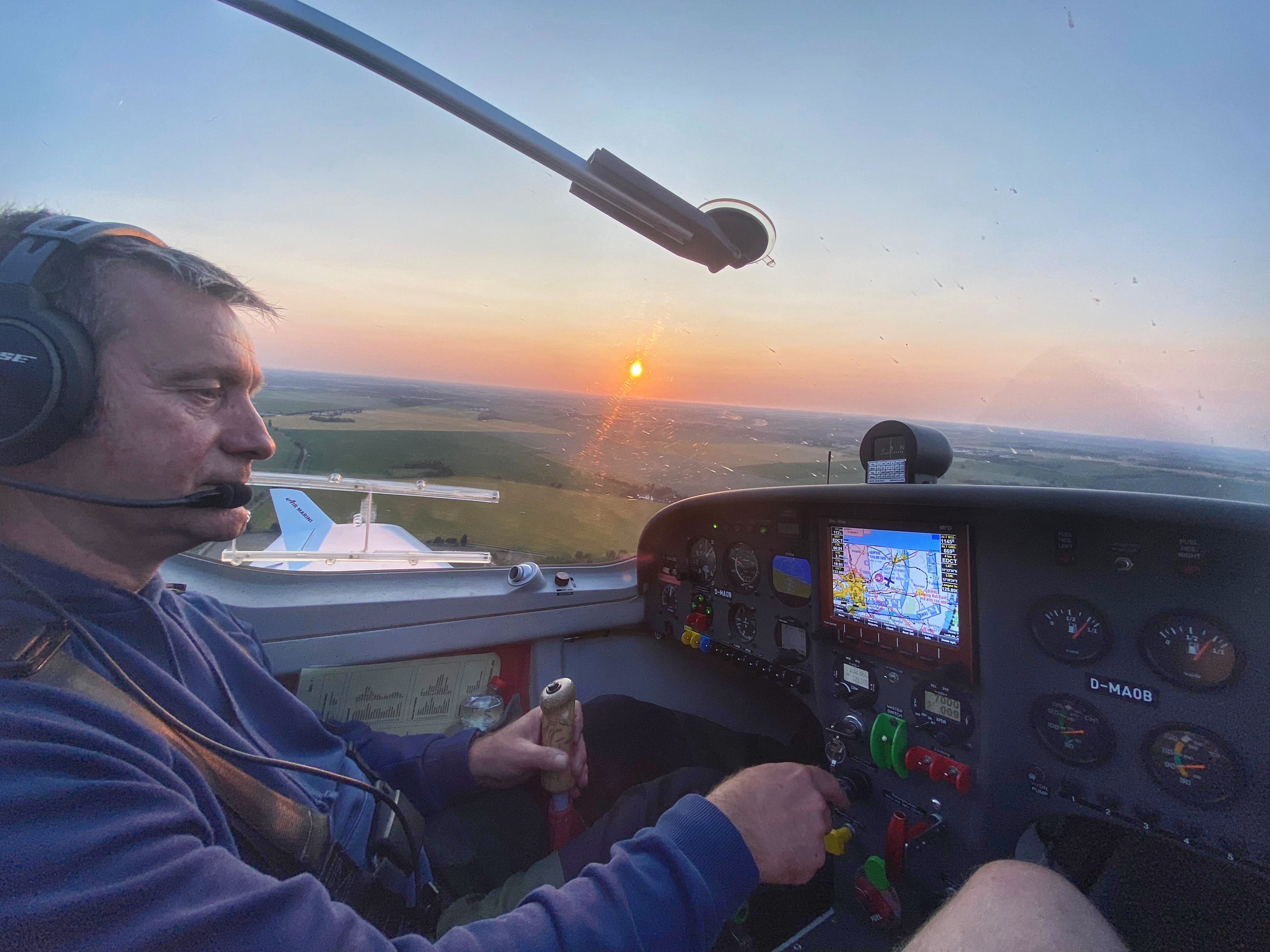 Ein Pilot steuert ein kleines Flugzeug bei Sonnenuntergang. Der Blick geht über das Cockpit auf die weite Landschaft darunter.
