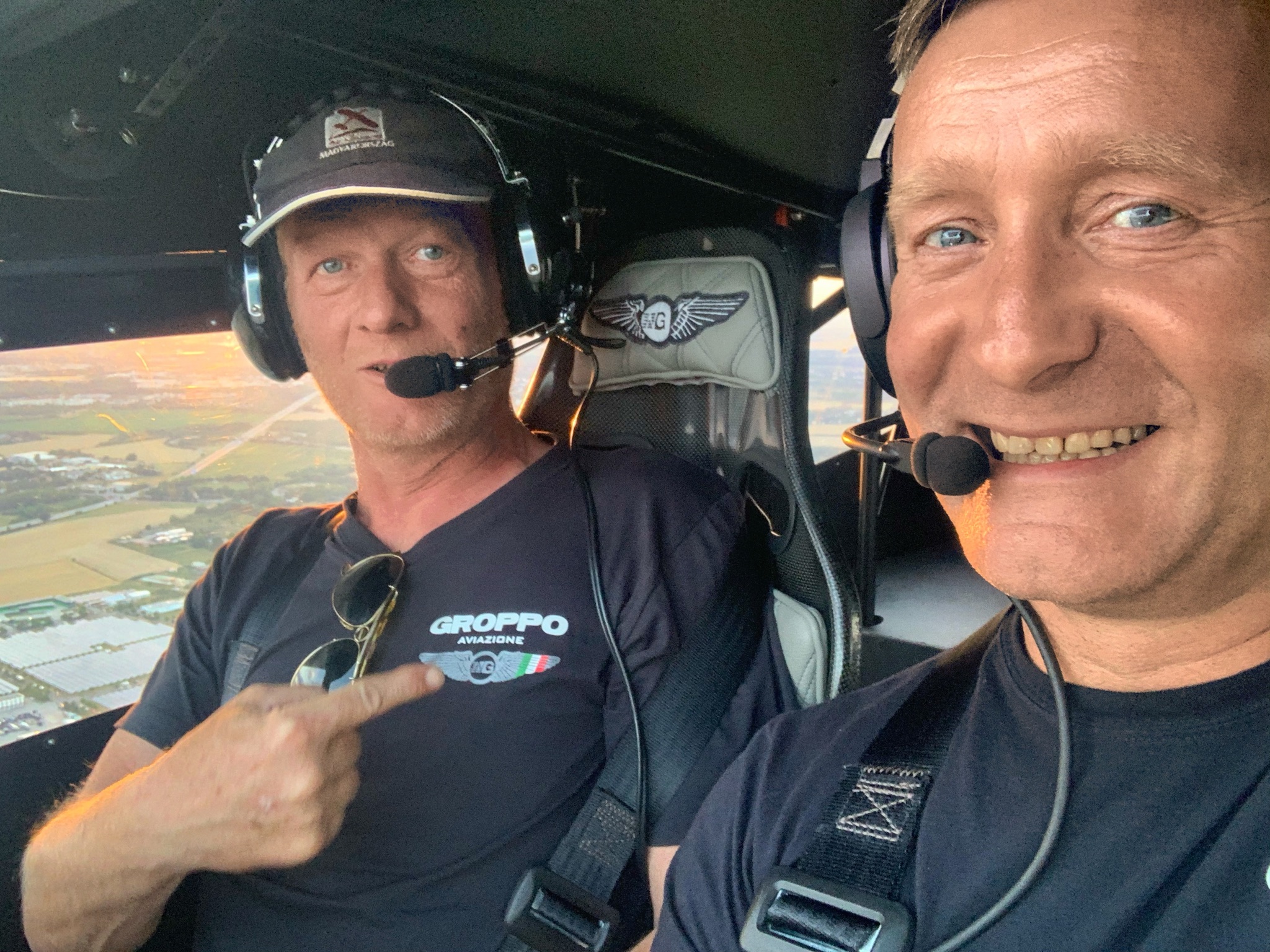 Zwei Männer im Cockpit eines Flugzeugs, beide tragen Headsets und lächeln in die Kamera, während sie während eines Flugs die Aussicht genießen.