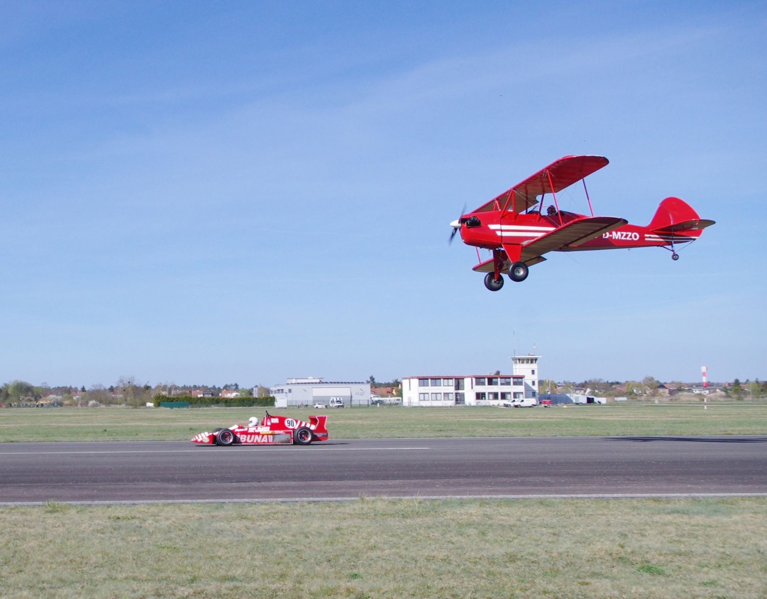 Ein rotes Doppeldecker-Flugzeug fliegt über einem Flugplatz, wo ein roter Rennwagen fährt. Im Hintergrund sind Gebäude und ein Turm zu sehen.