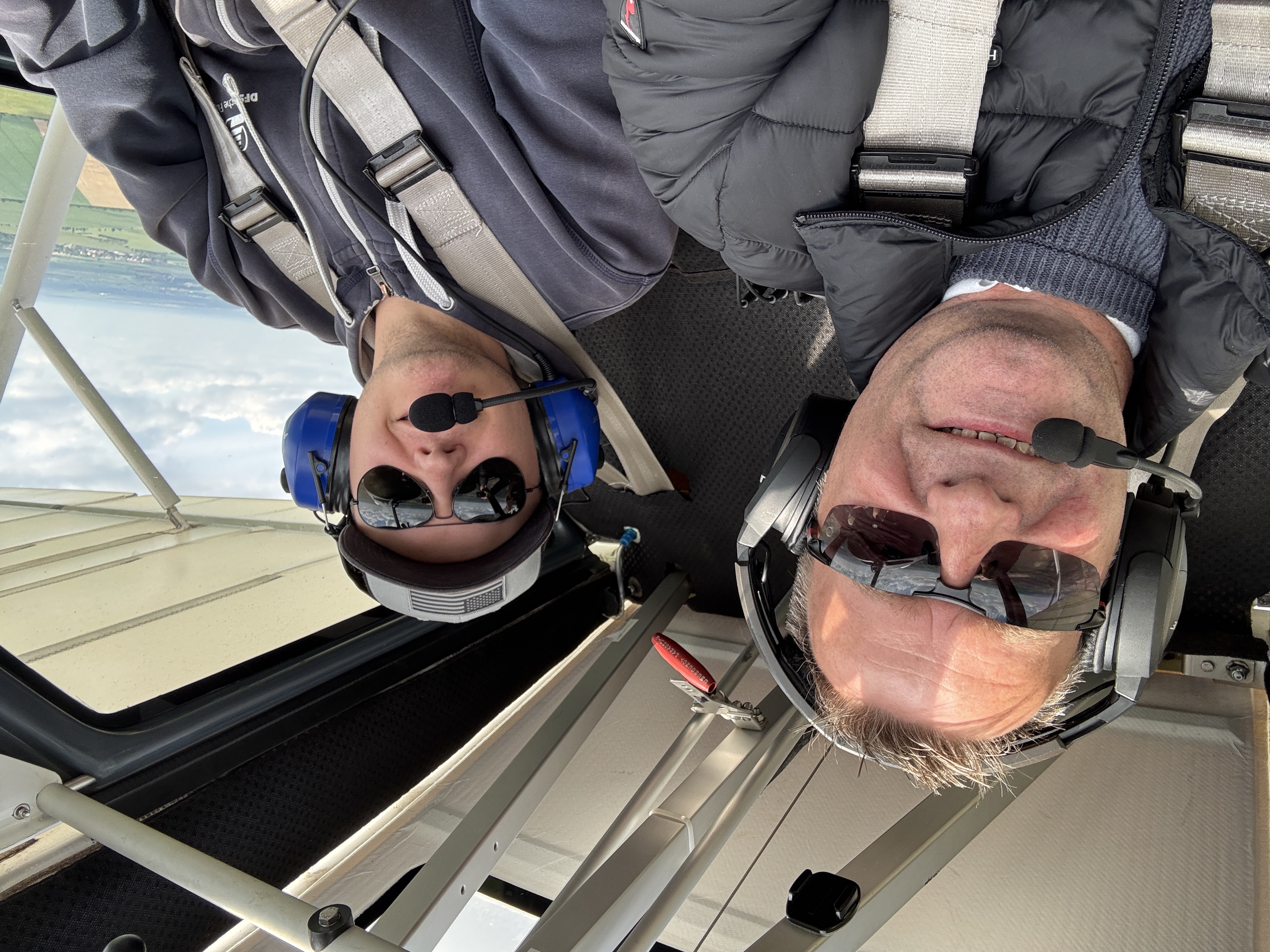 Zwei Männer mit Headsets sitzen im Cockpit eines kleinen Flugzeugs, während sie über eine Landschaft fliegen.
