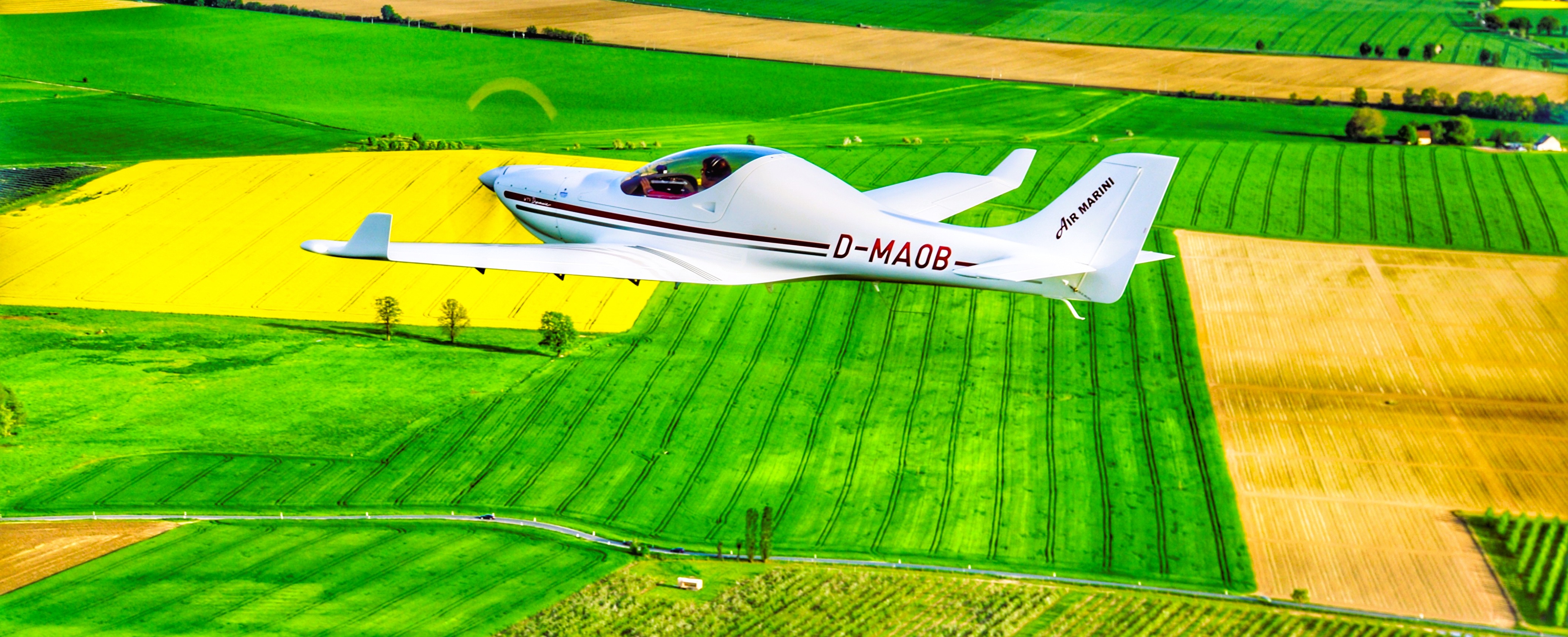 Ein Leichtflugzeug fliegt über eine malerische Landschaft mit grünen und gelben Feldern.