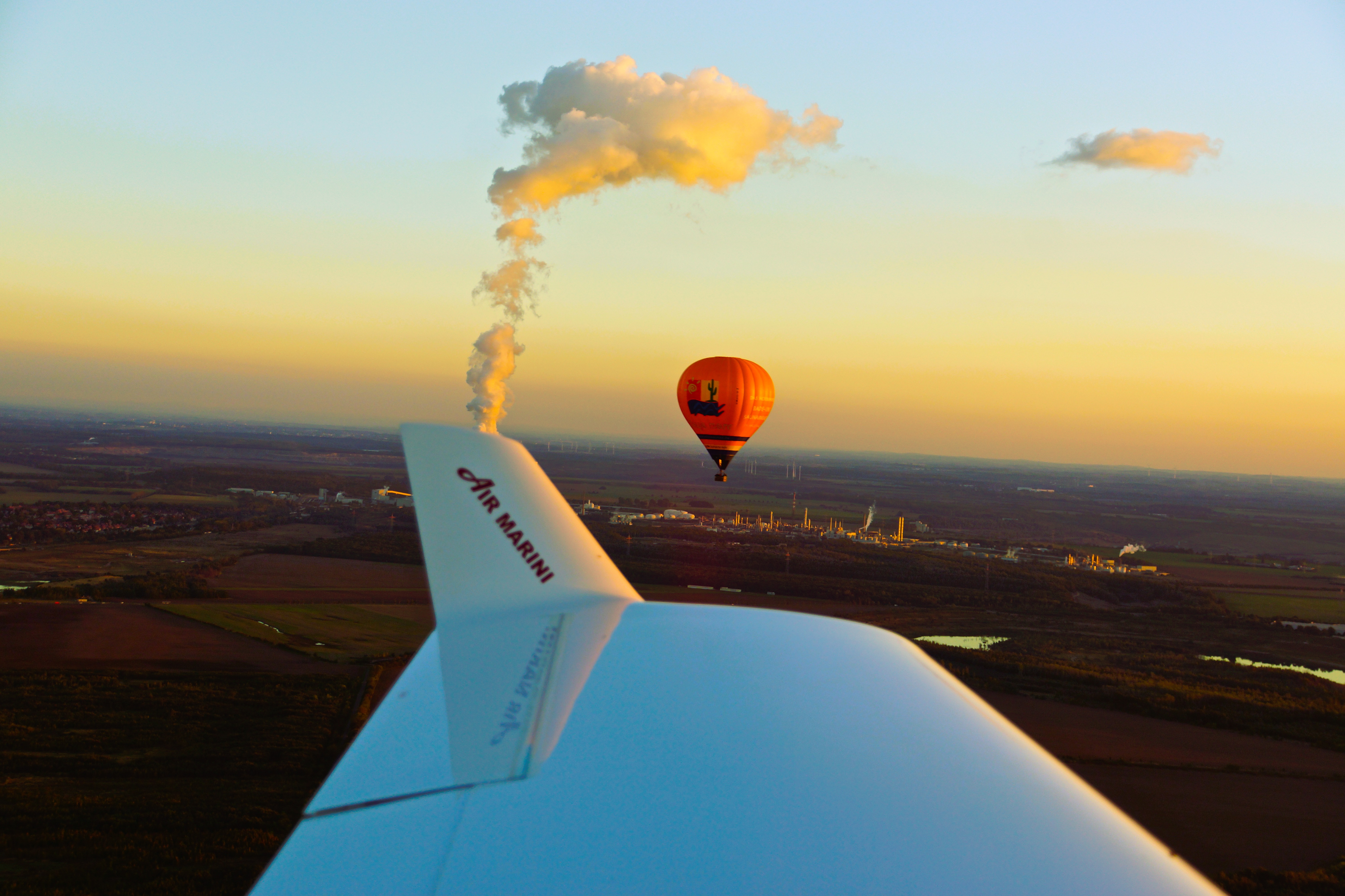 Ein Heißluftballon fliegt über eine Landschaft bei Sonnenuntergang, mit einem Flugzeugflügel der Marke Air Marini im Vordergrund und einer Fabrik im Hintergrund, aus der Rauch aufsteigt.