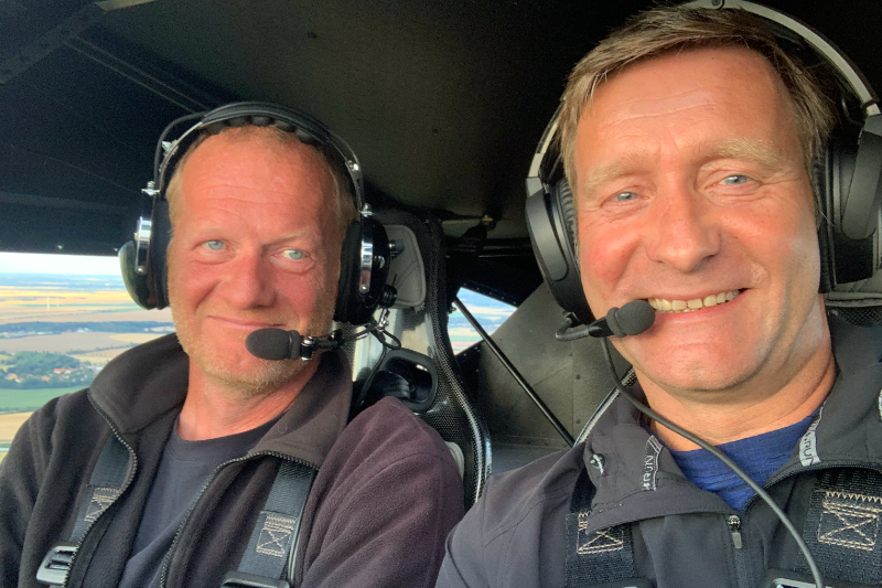 Zwei Männer mit Headsets lächeln in die Kamera, während sie im Cockpit eines Flugzeugs sitzen