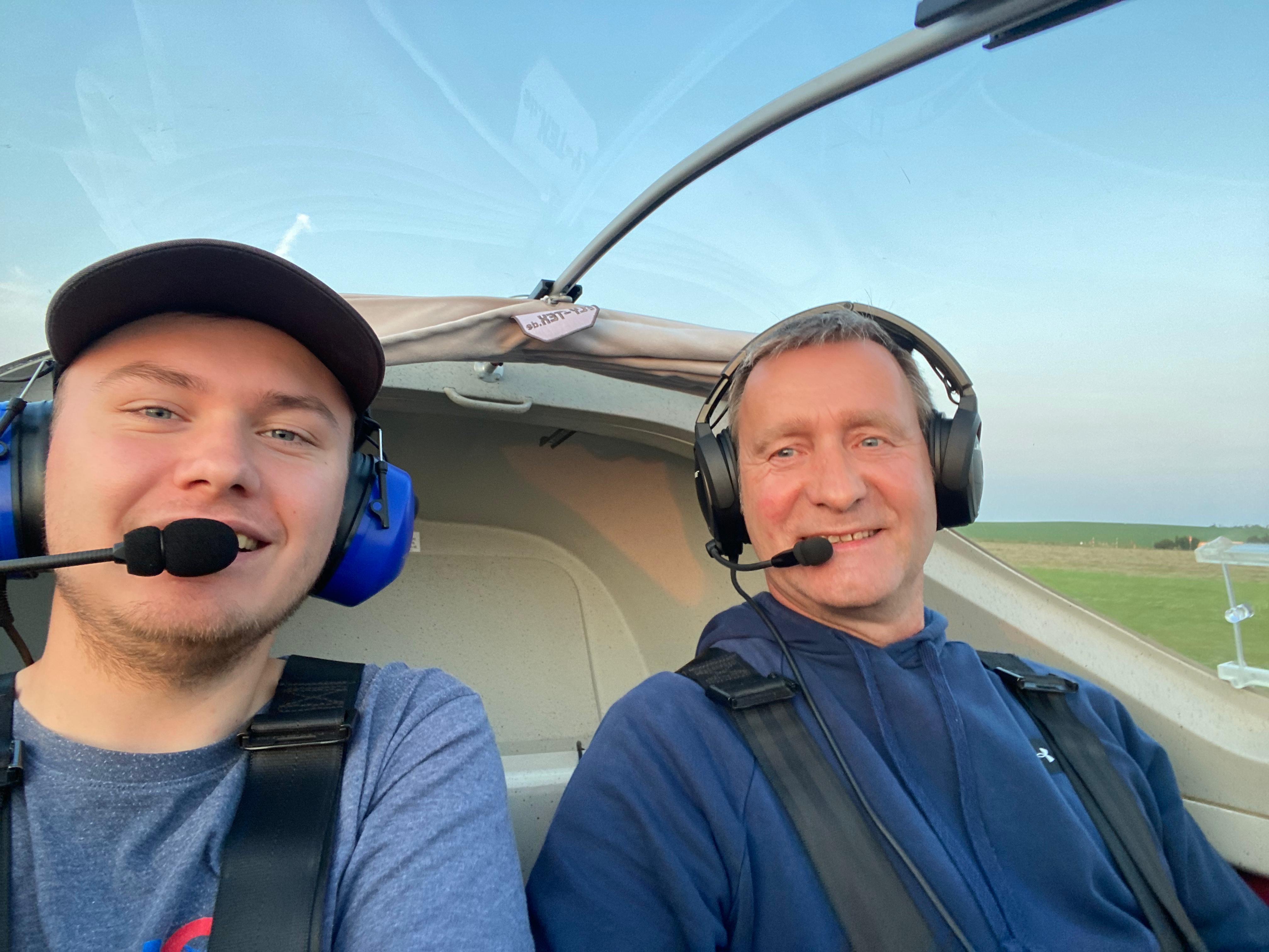Zwei Männer tragen Headsets und sitzen lächelnd in einem Flugzeug-Cockpit, während sie für ein Selfie posieren.