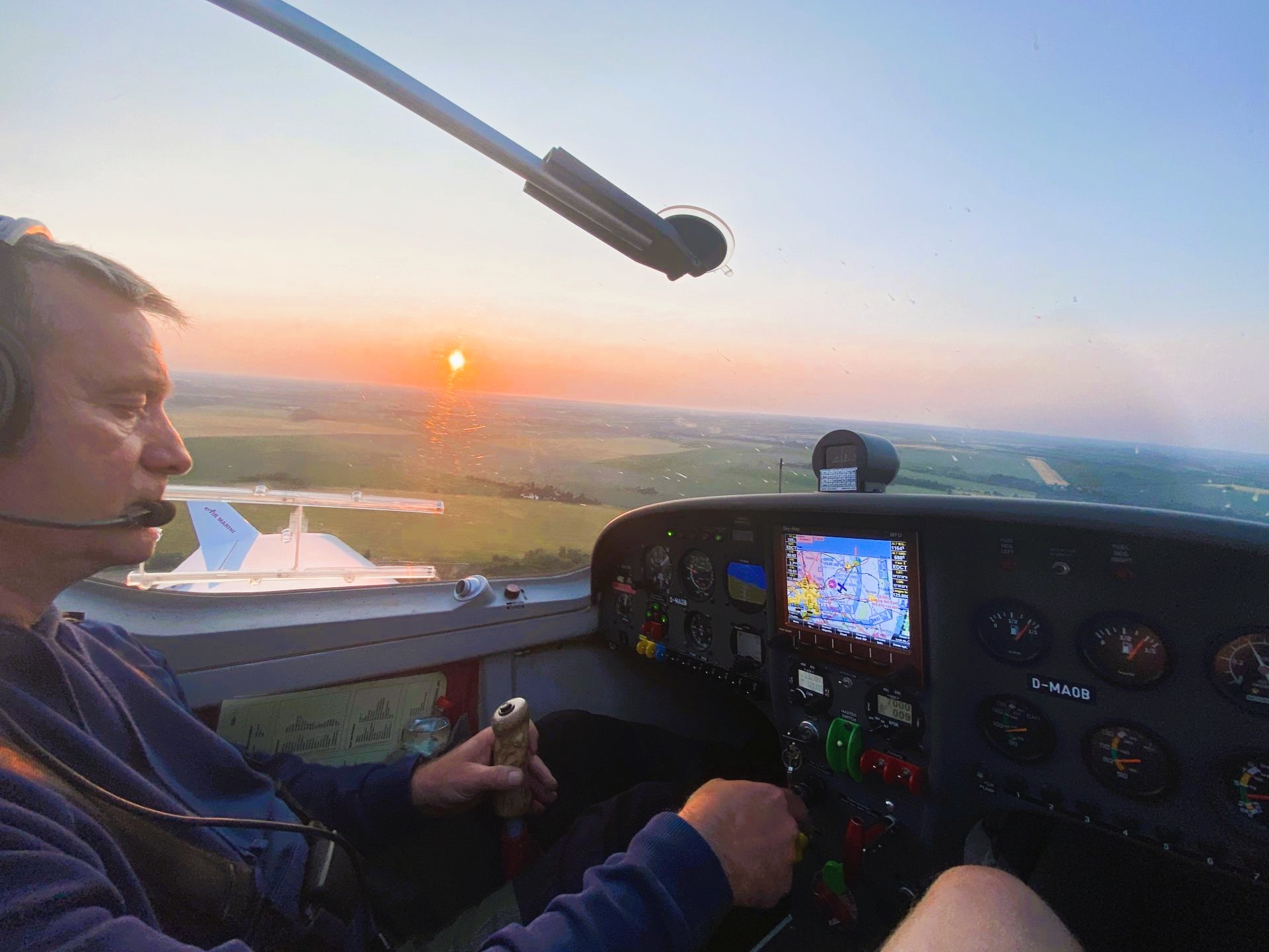 Ein Pilot fliegt ein kleines Flugzeug und genießt die Aussicht auf einen wunderschönen Sonnenuntergang über der Landschaft. Das Cockpit ist mit verschiedenen Instrumenten ausgestattet.
